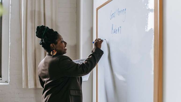 Imagem mostra uma professora, negra, com cabelos trançados em frente a um quadro