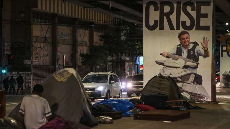 Em São Paulo, pessoas em situação de rua ocupam barracas em frente a um cartaz de protesto contra o presidente Jair Bolsonaro