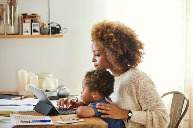 Boy looking at mother using digital tablet. Woman sitting with son at table in kitchen. She is working from home.