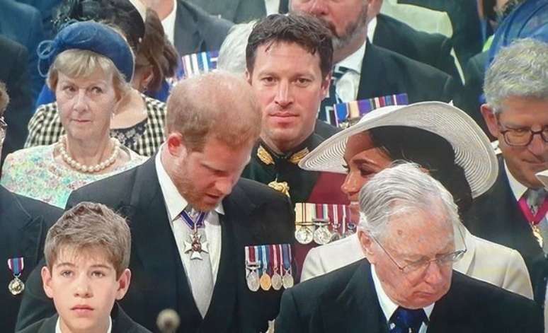 O militar discretamente a postos atrás do príncipe Harry e da duquesa Meghan