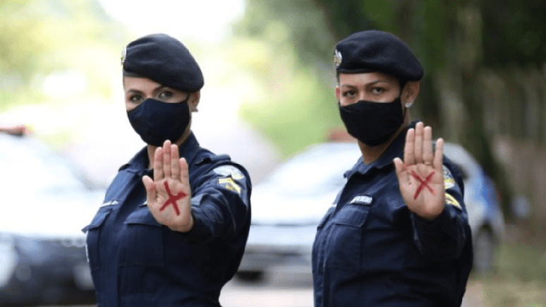 A foto mostra duas policiais com um x vermelho na mão, símbolo contra a violência doméstica