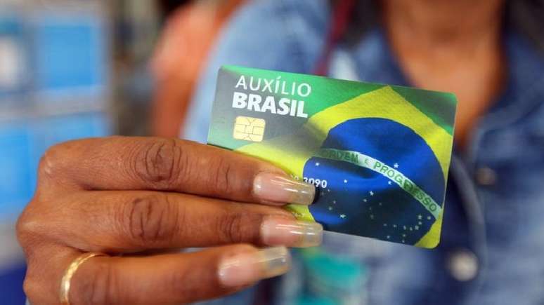 A foto mostra uma mulher negra segurando o cartão do Auxílio Brasil