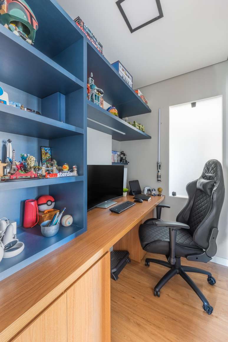Para o cliente que trabalha com videogames, a marcenaria adotou o MDF com visual amadeirado. J&aacute; no m&oacute;vel superior azul, os arm&aacute;rios abertos s&atilde;o perfeitos para expor a cole&ccedil;&atilde;o geek e, em geral, a parede de estantes permite amplo armazenamento e um visual especial ao espa&ccedil;o de trabalho| Projeto Bruno Moraes Arquitetura.