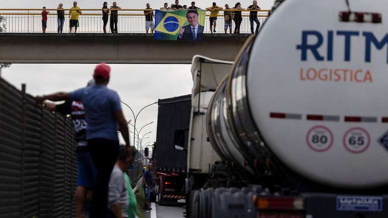 Estrada bloqueada em Jacareí (SP); ao fundo, em passarela, apoiadores do presidente seguram bandeira com foto de Bolsonaro