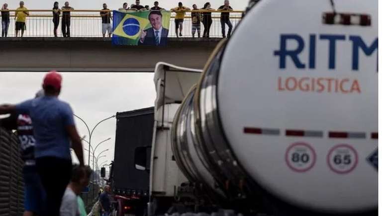 Estrada bloqueada em Jacareí (SP); ao fundo, em passarela, apoiadores do presidente seguram bandeira com foto de Bolsonaro