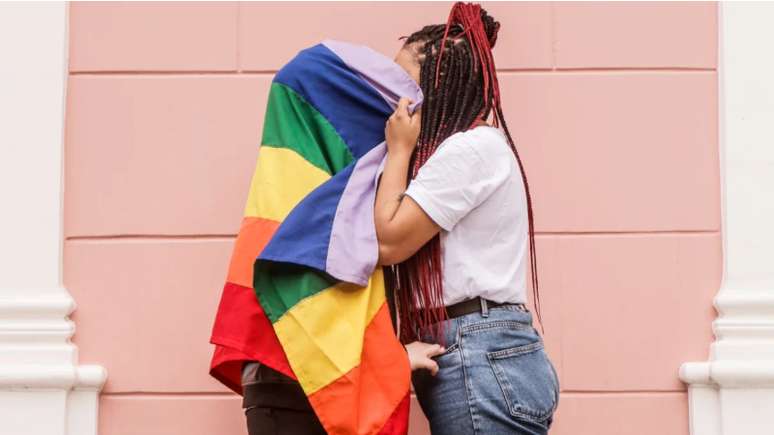 Imagem mostra casal de mulheres se beijando atrás de uma bandeira do arco-íris, símbolo do movimento LGBTQIAP+.