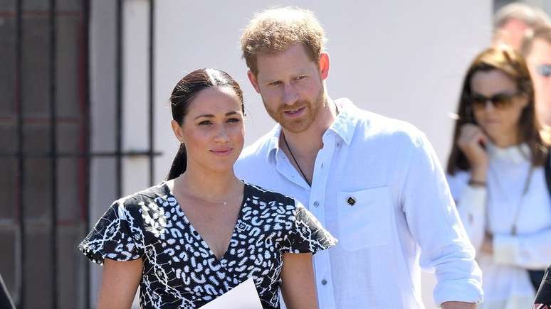 O príncipe Harry, usando seu cordão característico, ao lado de Meghan na África do Sul, meses antes de deixarem os deveres reais