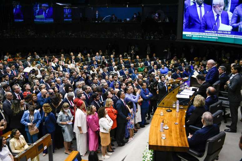 Lula toma posse no Plenário da Câmara dos Deputados. Lula lê um papel da mesa da Câmara enquanto uma multidão de pessoas em roupas sociais o observa do plenário