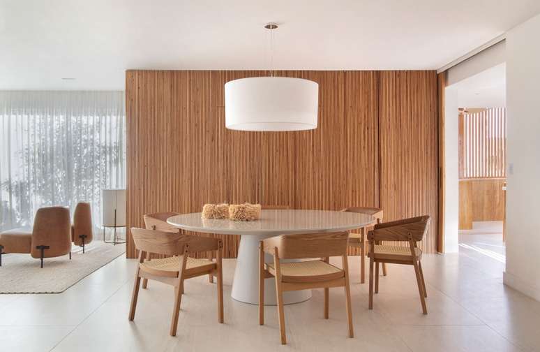 Sala de jantar minimalista com mesa redonda, lumin&aacute;ria, cadeiras de madeira e parede revestida com madeira ripada.