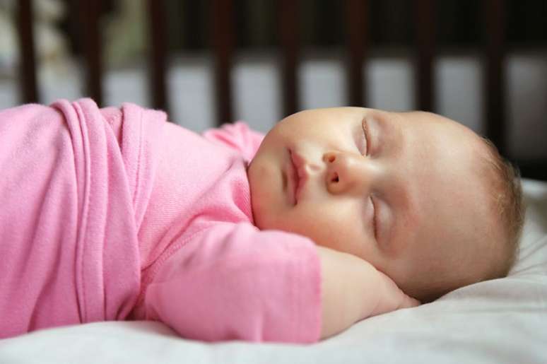 sleeping baby.  She is wearing a pink dress, has white skin and is lying on a mattress with a white sheet.  Lying on the stomach.