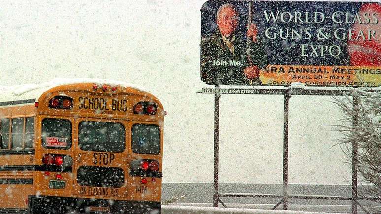 Um ônibus escolar passa por um outdoor promovendo a convenção anual da NRA em 1999, após o massacre de Columbine