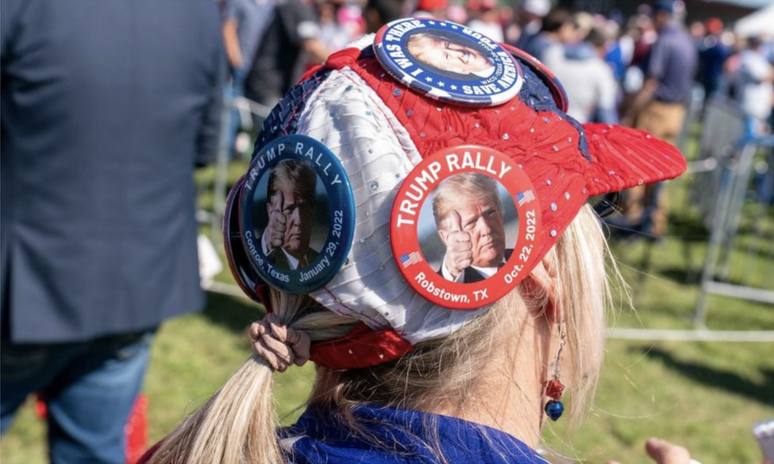 Mulher de costas usando boné com broches de apoio a Trump em comício