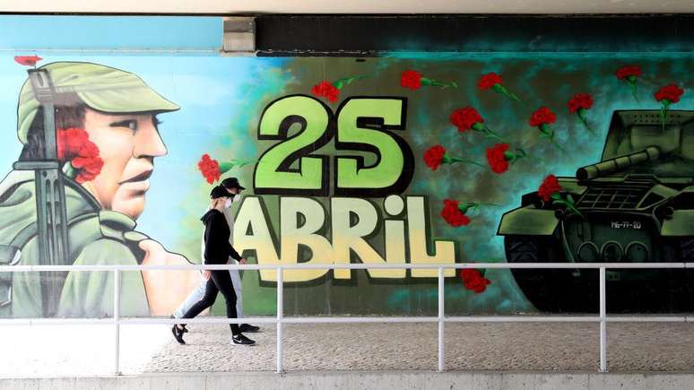 Duas pessoas caminhando em frente a mural em homenagem a 25 de abril, com militar usando cravo 