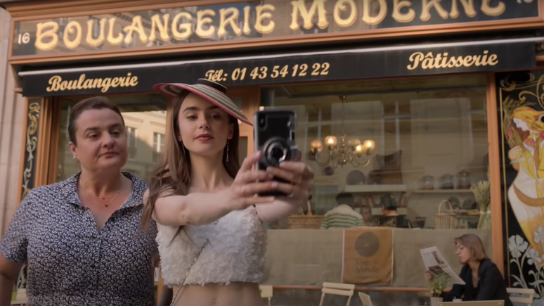 Emily (Lily Collins) tirando uma selfie com a atendente da padaria em cena de Emily em Paris.