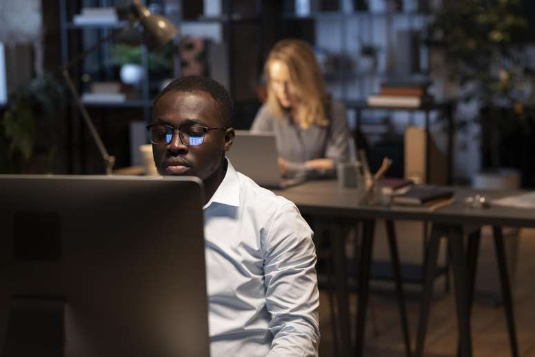Um homem negro olha para a tela de um computador, que aparece refletida na lente dos óculos dele. Ele tem cabelo raspado e camisa branca. Atrás e desfocada, uma mulher loira trabalha num laptop.
