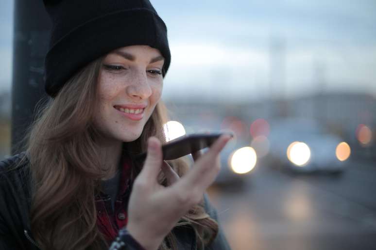 Uma jovem sorridente segura um celular na horizontal próximo a boca. Ela tem a pele alva e sardinhas no rosto. Usa gorro e casaco pretos. Atrás e desfocados, há carros com faróis acesos.