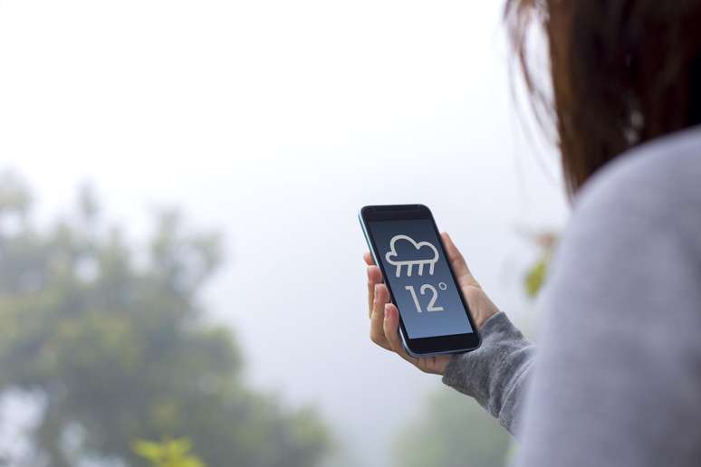 Mão de mulher ao ar livre segurando celular, no qual aparece previsão de chuva na tela