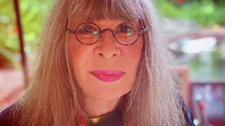 Foto aproximada do rosto de Rita, que sorri contidamente, em &aacute;rea verde