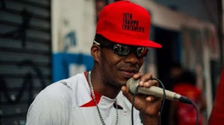 MC Cesinha, artista agredido duas vezes na Cracolândia, usa uma camiseta branca, boné vermelho e segura um microfone.