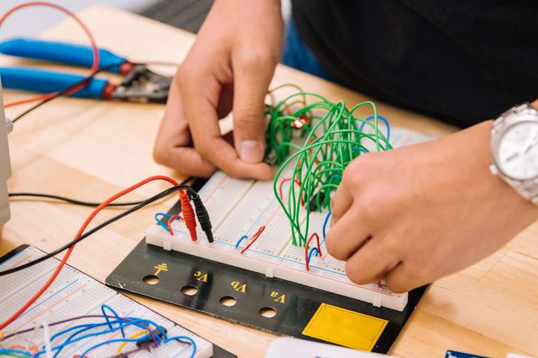 Mãos trabalham em uma placa de protótipo com fios finos azuis, vermelhos e verdes. Ela está apoiada numa bancada de madeira. A placa plástica é quadrada, branca e tem centenas de micro furos onde os fios estão conectados.