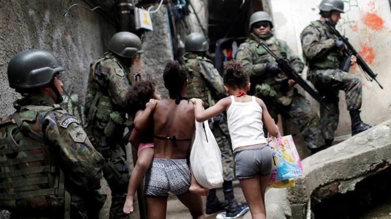 Imagem mostra uma mulher e duas crianças negras de costas passando por uma rua cercada por policiais.