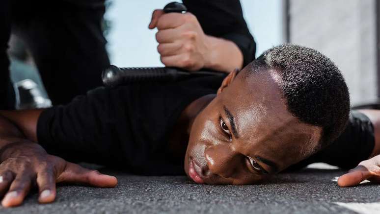 Imagem mostra homem negro no chão, sendo agredido e imobilizado por policial.
