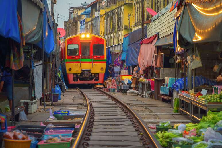 Estrada de ferro tailandesa com um trem local funciona através do mercado de Mae Klong na província de Samut Songkhram, Tailândia. 