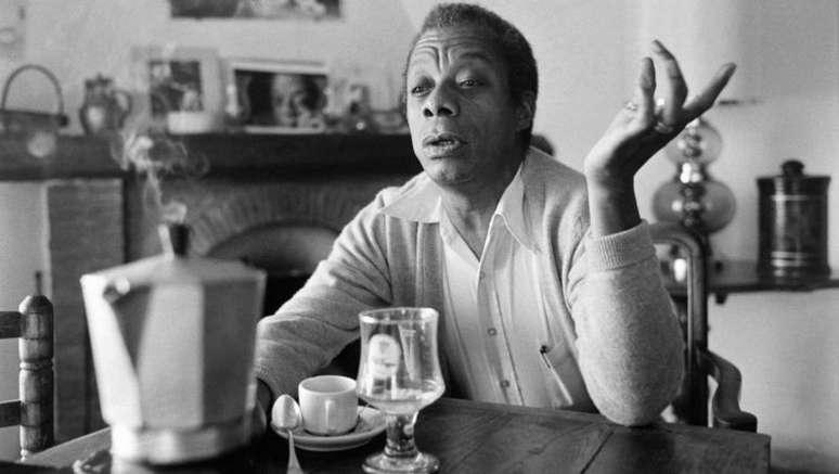 Imagem em preto e branco mostra James Baldwin, um homem negro de meia idade sentado e tomando um café.