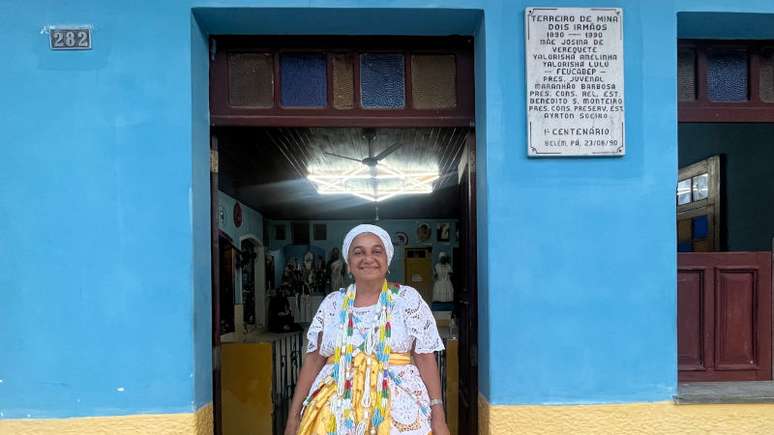 Imagem mostra a ialorixá mãe Eloísa de Badé, zeladora do terreiro. Ela está com vestimentas brancas e na entrada do espaço.