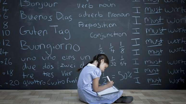 Menina escrevendo em caderno em frente a lousa com palavras em várias línguas