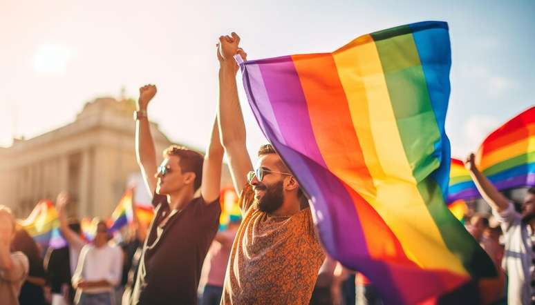 Foto mostra 2 homens em primeiro plano, onde um deles segura a bandeira que representa a luta das pessoas LGBTQIAPN+, durante um evento, com várias outras pessoas desfocadas em segundo plano