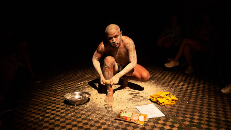 Foto mostra uma fotografia da Série “Até os filhos do urubu nascem brancos”, de Waleff Dias. Na imagem, há um homem abaixado, esfregando farinha em seu corpo já coberto pela textura esbranquiçada.