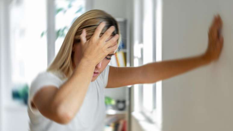 Mulher loira vestindo blusa branca aparenta sofrer com tontura; ela está com uma mão na cabeça e a outra na parede, e o fundo da foto está borrado