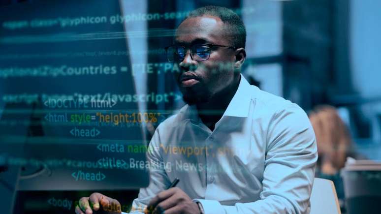 Imagem mostra um homem negro, careca, com óculos e camisa social branca olhando para a tela de seu computador enquanto digita. O homem está em uma espécie de escritório. Na foto existe um filtro com códigos de programação, simulando como o racismo algorítmico pode ser reproduzido