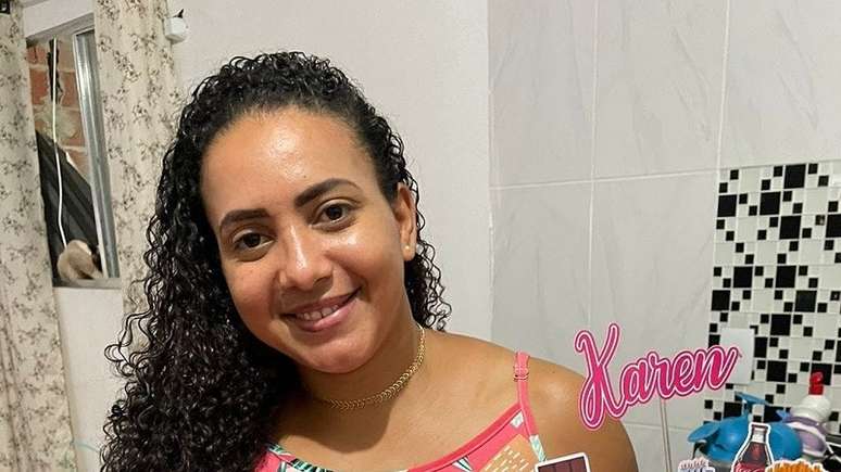 Imagem mostra Karen Cristina, uma mulher negra de pele clara e de cabelos cacheados e pretos longos. Ela está dentro de casa, veste uma blusa rosa e segura um bolo de aniversário.