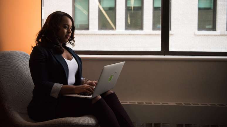 Imagem mostra uma mulher negra sentada em uma poltrona e usando um notebook.