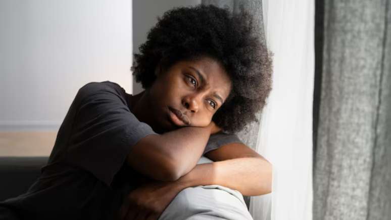 Imagem mostra um adolescente negro debruçado em um sofá, olhando para o lado de fora da janela com expressão triste.