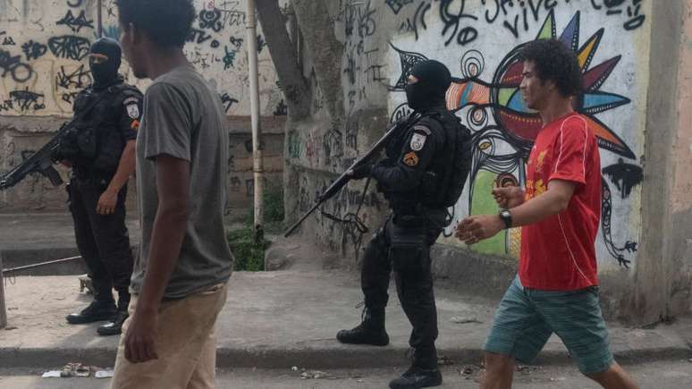 Imagem mostra dois homens negros caminhando na rua e dois policiais armados.