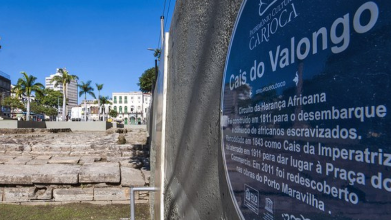 À direita se encontra uma placa pregada à parede com informações sobre o Cais do Valongo e ao fundo o patrimônio histórico