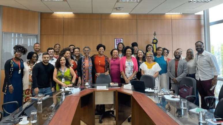 25 pessoas em pé a frente de uma mesa de vidro com detalhes em madeira