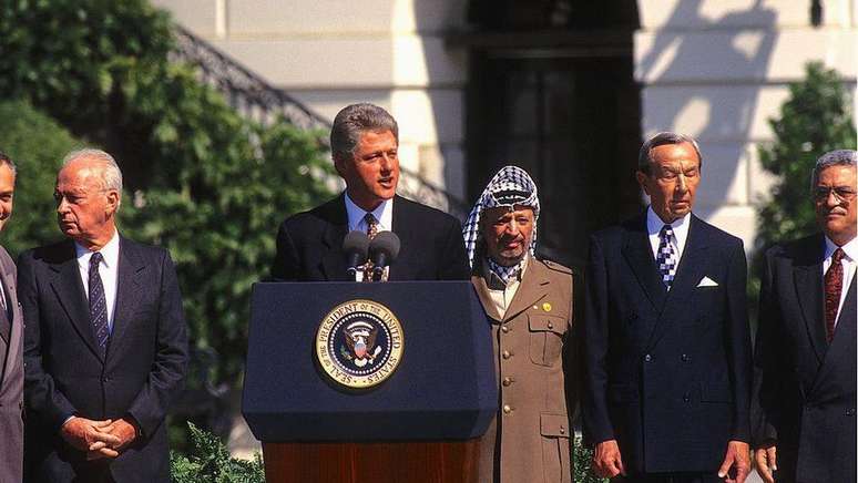 O primeiro-ministro de Israel, Yitzhak Rabin, e o l&iacute;der da OLP, Yasser Arafat, ao lado do ent&atilde;o presidente dos EUA, Bill Clinton, na assinatura dos acordos; Mahmoud Abbas (&agrave; direita) tinha 57 anos na &eacute;poca.