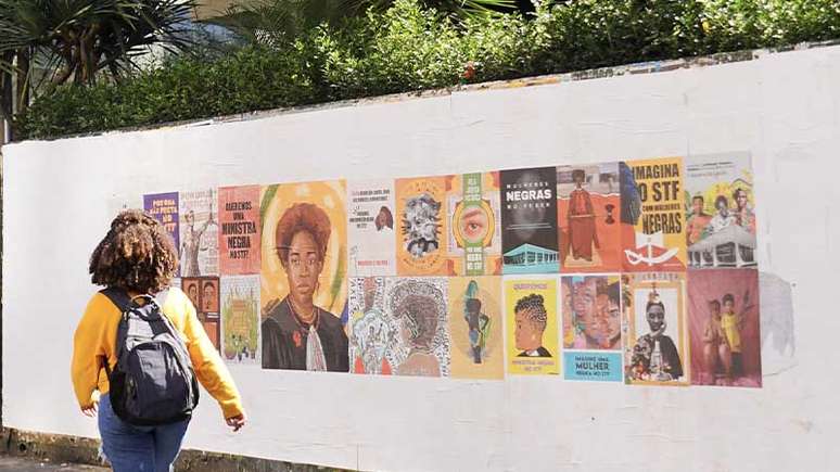 Mulher negra, de costas passando ao lado do mural a céu aberto, com 24 obras, pedindo uma ministra do STF negra.