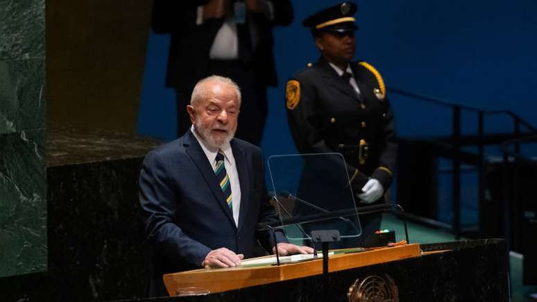 A imagem mostra o presidente Luiz Inácio Lula da Silva discursando. Ele é um grisalho, com barba grisalha, usando um terno. Ao seu fundo, uma mulher negra com farda fazendo a vigilância do presidente. A sua presença alude à fala do presidente sobre igualdade racial.