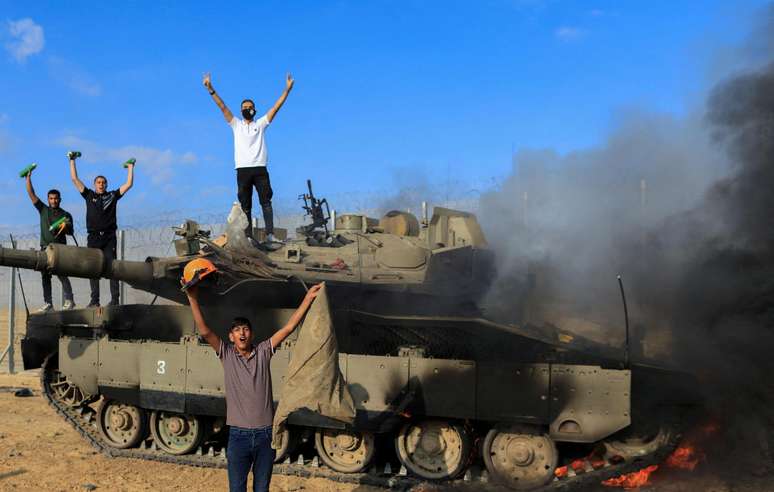 Palestinos comemoram enquanto um veículo militar israelense pega fogo depois de ter sido atingido por homens armados que se infiltraram em áreas do sul de Israel