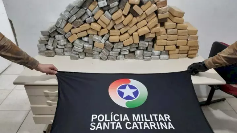 Policiais seguram bandeira da PMSC; ao fundo, maconha apreendida está colocada sobre escrivaninha