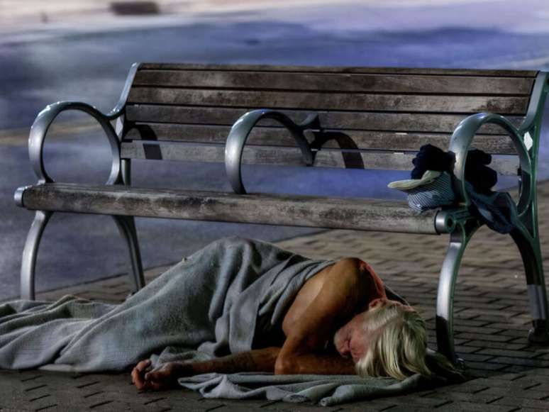 Imagem mostra homem em situação de rua deitado no chão, embaixo de um banco de praça.