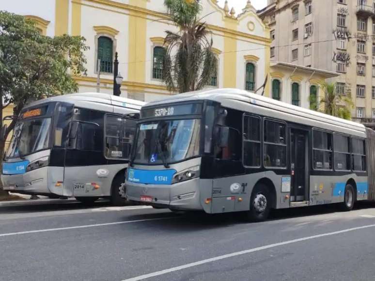 Imagem mostra doiz ônibus cinza e azul em rua de São Paulo. Ao fundo há um prédio antigo, nas cores branco e amarelo.