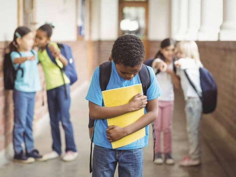 Imagem mostra um menino negro com mochila nas costas e livros nas mãos e outras crianças apontam para ele.