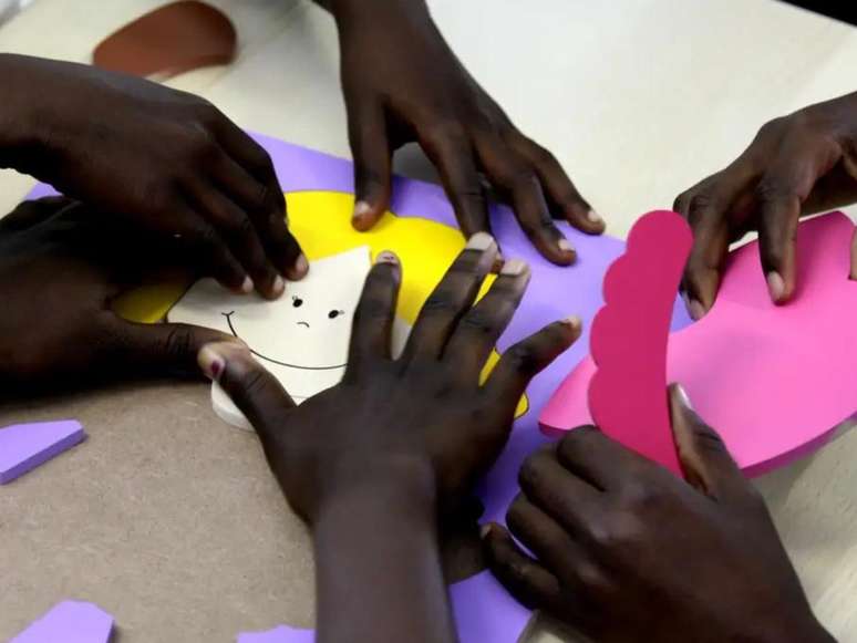 A imagem mostra mãos de crianças negras na escola fazendo uma atividade com papéis