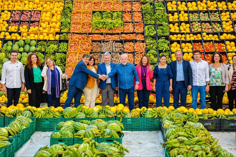 Grupo de pessoas que inclui o presidente Lula, ministros e o prefeito do Recife, João Campos, estão em um cenário repleto de caixas de frutas e legumes